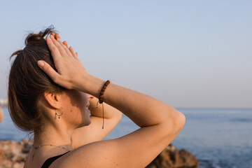 A young Caucasian woman with brown hair stands on rocky beach, gazing at the sea. The sun sets in...