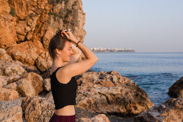 A young Caucasian woman with brown hair stands on rocky beach, gazing at the sea. The sun sets in the background, casting warm light on the scene.