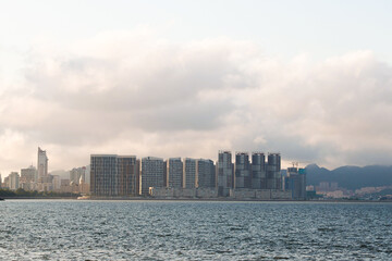 Naklejka premium Stunning Waterfront Skyline with Modern Buildings Amidst Clouds and Mountains A Serene Urban Landscape Perfect for City Life Enthusiasts and Nature Lovers Alike