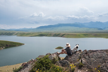 A woman and her dog friend sit on rocks above a mountain lake in summer, admiring the surrounding landscape of Kazakhstan's wild nature. Bestyubinskoye Reservoir, Bestobe on the Charyn River.