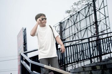 A man dressed in a simple white t-shirt and sunglasses stands confidently on an outdoor stairway, capturing a casual yet stylish scene with an urban backdrop.