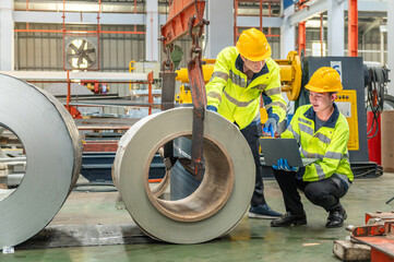 Two industrial factory workers in yellow safety helmets and reflective vests inspecting large steel...