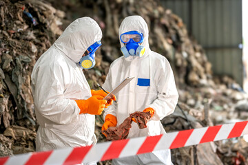 Two workers in full protective hazmat suits with blue goggles and orange gloves examining a sample or specimen at a hazardous waste site with rocky background and safety barrier