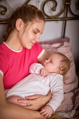 Young woman with light brown hair and caucasian features lovingly cradles sleeping infant girl in pink and white attire, evoking warmth and tenderness