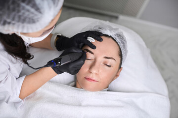 Professional performs eyebrow permanent makeup procedure on relaxed adult woman in clean, tranquil...