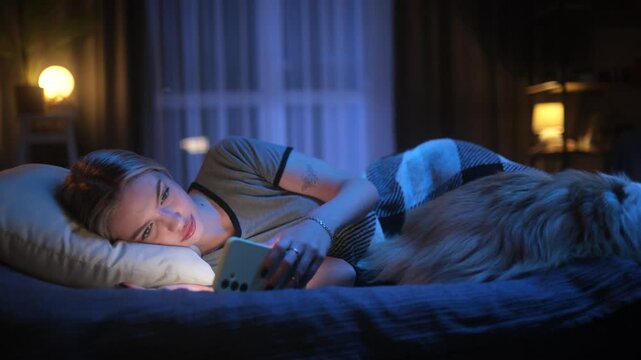 Young woman scrolling on her smartphone in bed at night, yawning, and falling asleep with her dog beside her. Cozy evening routine