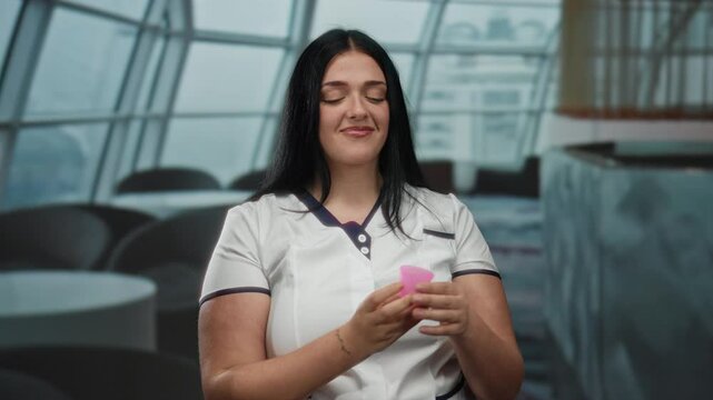 Young woman smiling holding pink menstrual cup indoors giving thumbs up in modern setting showcasing feminine empowerment and environmental awareness with confidence.