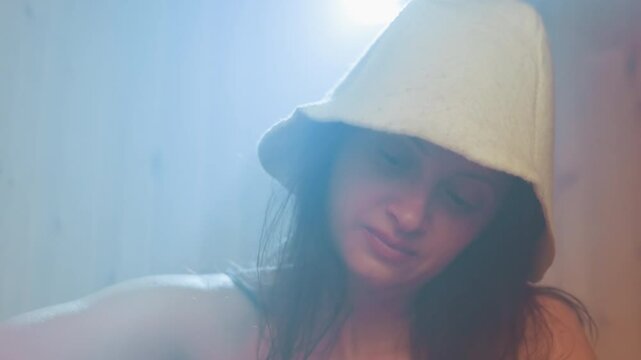 misty spa scene with young woman wearing felt spa hat illuminated by soft backlight gently massaging cream into leg during serene self care ritual in wooden steam cabin ambience