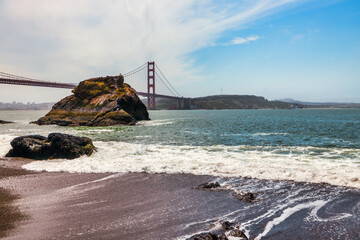 Golden Gate Bridge view from the rocky shoreline on a clear day in San Francisco, California. Coastal waves, iconic red architecture, and Sutro Tower in the distance capture the essence of the Bay