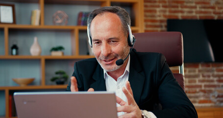 Mature businessman CEO holding video conference call with clients, wearing wireless headphones with microphone. Handsome worker having video call on laptop computer.