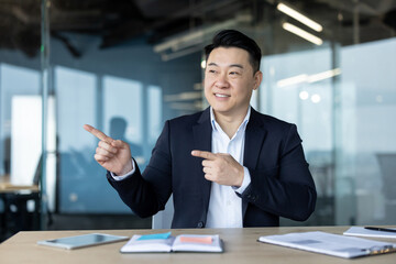 Smiling young Asian male office worker sitting at his desk and pointing with his fingers to the...