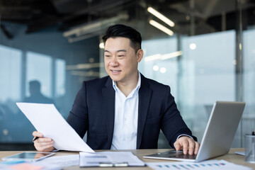 Smiling young Asian male businessman sitting in the office at the table and working on a laptop and...