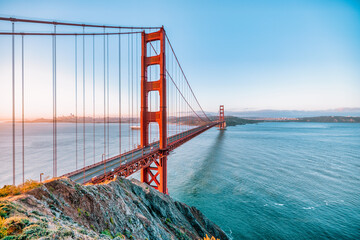 Golden Gate Bridge at sunrise in San Francisco, California. Scenic view from Battery Spencer with soft pastel sky, calm bay waters, and iconic red architecture. Tranquil travel landscape.