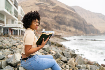 Young woman reading a book on a rocky beach, enjoying the ocean view © Guillermo Spelucin