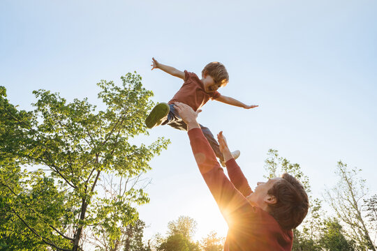Father tossing his laughing joyful son into the air against a bright sky in a park on a sunny day. Cheerful boy playing with his dad outdoors, creating happy childhood memories together. Copy space