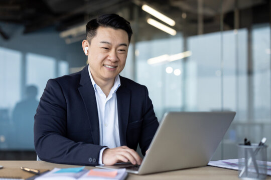 Asian smiling young businessman in headphones and suit sitting in the office at a desk and working on a laptop