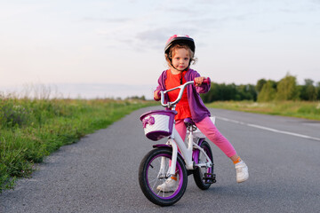 A determined little girl in a helmet stands astride her small bicycle on a paved road, looking...