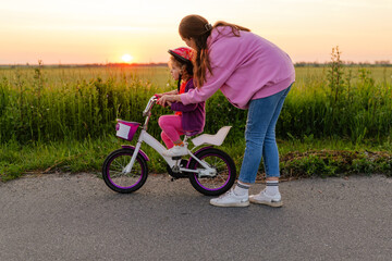 A mother helps her young daughter learn to ride a bicycle on a paved road, with a golden sunset...