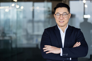 Portrait of a young Asian man wearing glasses and a suit standing in an office lobby, arms crossed...