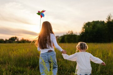 Two young girls, holding hands, walk away into a sunlit field with a colorful pinwheel, as the sun...