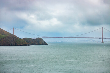 Close-up view of the Golden Gate Bridge rising behind rugged coastal cliffs of Marin Headlands, captured on a moody overcast day in San Francisco Bay, California