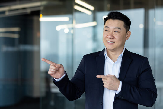 Asian smiling young businessman standing in office wearing suit and pointing with fingers to the side at empty space, presenting and advertising