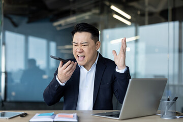 An angry Asian man sits in a chair at his desk and shouts into the loudspeaker of the mobile phone...