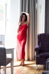 Full-length portrait of stylish brunette model in long red gown walking upstairs and posing near...