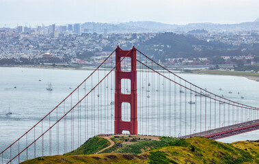 Panoramic view of the Golden Gate Bridge spanning San Francisco Bay with cityscape, lush hills, and boats, captured from a high vantage point on a cloudy spring day