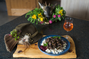 happy hunting of a young roe buck - fresh innards from a roe buck hunt, roasted with herbs, rice and cranberries, served on a blue plate and a glass of wine, decorated with the hunting hat