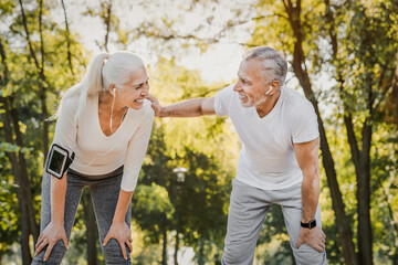Active hobby on retirement. Grey haired caucasian man with beautiful wife strolling at summer park and smiling cheerfully. Fitness retired couple in sport attire taking break after morning run.