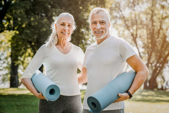 Lovely senior man and old woman holding yoga mat for exercising outdoor. Aged husband and wife ready for yoga at park. Active healthy retired couple looking at camera before a postural session.