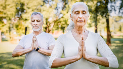 Senior man and woman retired family people standing in park with their hands together closing eyes, enjoying the tranquility of practicing yoga class together. Active sport rehabilitation concept
