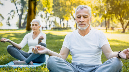 Yoga practice at park. Peaceful grey haired man and woman sitting in lotus pose with closed eyes and meditating on fresh air. Couple on retirement keeping hands in mudra gesture and feeling calmness.