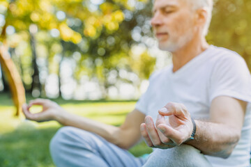 Yoga practice at park. Peaceful grey haired man sitting in lotus pose with closed eyes and meditating on fresh air. Retirement person keeping hands in mudra gesture and feeling calmness.