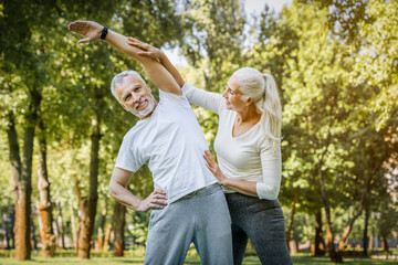 Senior fitness instructor is helping an elderly man stretching warming up during an outdoor workout...
