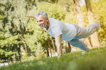 Side view shot of happy senior man in sportswear doing push ups on lawn in park sport exercise plank yoga workout outside. Fitness healthy lifestyle active retired people hobby rehabilitation concept