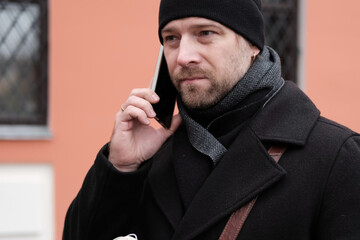 Handsome man wearing black coat and brown leather bag walking down on the city with mobile phone in...