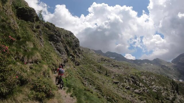 Two cyclists follow steep ridgeline path, rocky steps break narrow trail, layered peaks fade into distant haze.