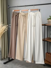 Three pairs of trousers hanging on a rack in different colors with a shelf and bag in the background