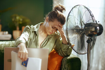 A distressed woman, hand on forehead with eyes closed, battles the combined discomfort of summer heat and the overwhelming burden of financial responsibilities, indicated by bills and a fan.