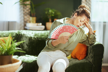 A middle-aged woman on a velvet couch struggles with intense summer heat, fanning herself with a...