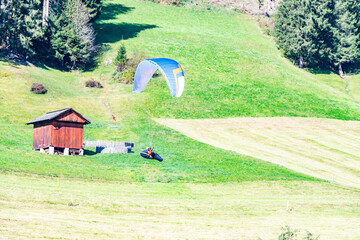 Dynamic shot of paragliding in the Italian Alps. A vibrant paraglider hovers over a green meadow next to a rustic wooden cabin. Adventure sport in nature.