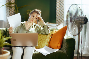 A distressed woman in her home office, fanning herself with papers and an exasperated hand on her forehead, struggles with heat and overwhelming work, beside a metallic fan.