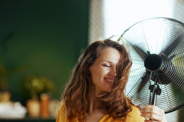 A woman with brown wavy hair smiles peacefully with closed eyes, embracing the cool breeze from a metal fan. This indoor scene depicts refreshing relief and comfort from the summer heat.