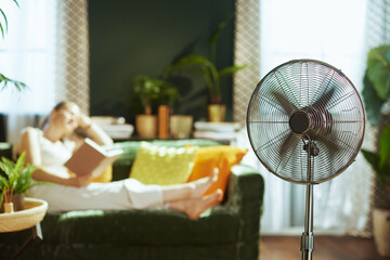 A young adult woman enjoys a book on a dark green velvet sofa in a cozy living room, complemented by a silver pedestal fan providing a cool breeze, creating an atmosphere of leisure and indoor comfort