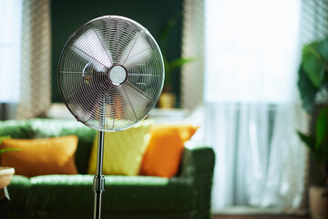 A prominent silver standing fan with spinning blades brings comfort to a modern living room, featuring a green sofa, sunlit windows with sheer white curtains, ideal for staying cool in warm weather.