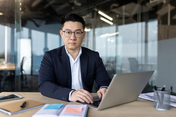 Fototapeta premium Portrait of a serious young Asian businessman sitting in the office at his desk, working on a laptop and looking at the camera