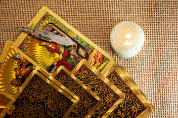 Tarot cards with vivid illustrations lie on burlap, with a quartz pendulum and a lit candle. A...