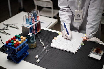 Unrecognizable Caucasian woman wearing lab coat and gloves recording scientific data in notebook...